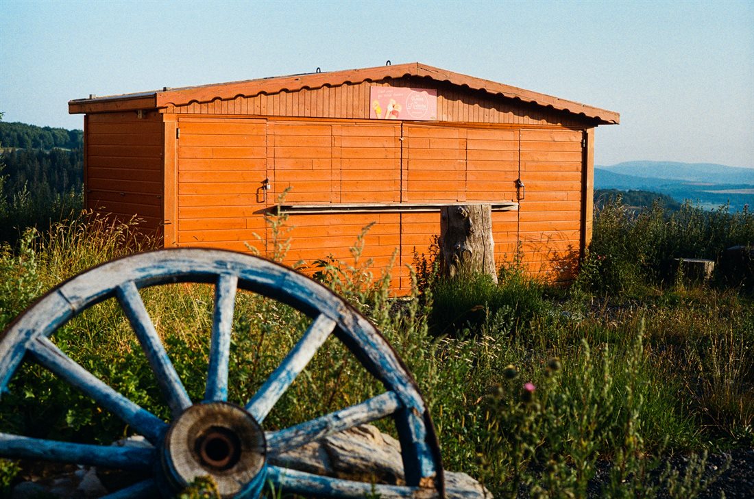 cabane en bois orangée, vente de glaces tourisme Gerbier-de-Jonc, Ardèche, roue de charrette bleue en premier plan, lieu herbu, en fond montagnes